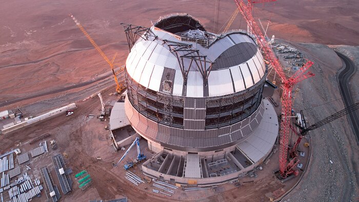 At the centre of the drone image, the ELT construction site stands out against the red sand of the Atacama desert. Its giant dome is covered in shiny metal corrugated plates, with part of the roof still open to the elements, and giant cranes surrounding the building. Halfway up, the building's steel frame is still showing where it is not yet covered by plates.