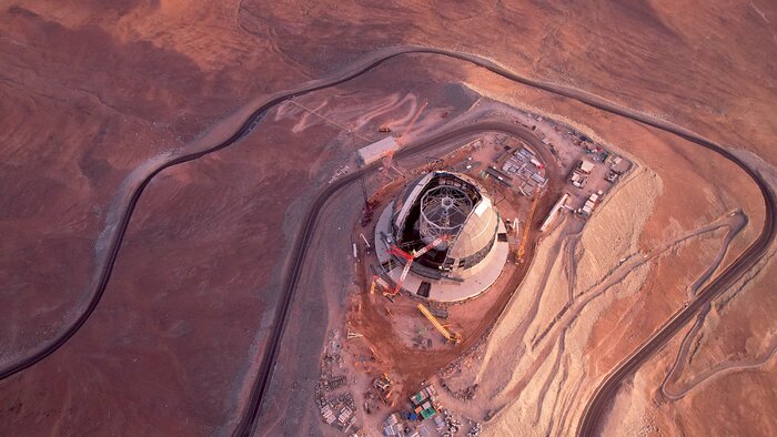 This aerial photo shows the entirety of Cerro Armazones, together with a serpentuous road that climbs up to the Extremely Large Telescope. The telescope is seen here in its entirety, still under construction, surrounded by cranes, containers and different vehicles. The landscape of the Atacama Desert is of a striking red-to-orange colour, while the sections that have been excavated have a pale brown colour.