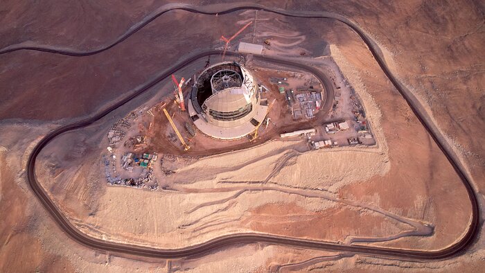 In this drone shot, taken from high up in the air, we look down at a road that carves itself up around Cerro Armazones, all the way to the top of the mountain, where the building stands that will house the Extremely Large Telescope. This building is at the centre of the image. Materials, cranes and vehicles look tiny. The road contrasts with the backdrop of the barren red mountain. The building appears to have the same sandy colour as the dug up sand.