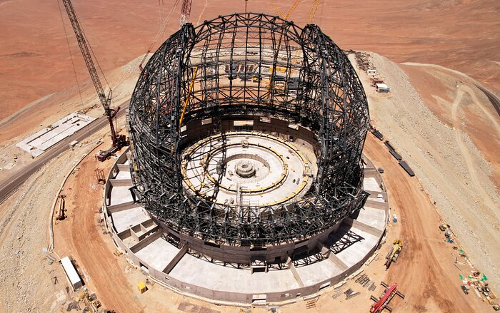 The grey steel skeleton of the ELT dome dominates this aerial image of the construction site. The hemispherical structure is atop a concrete base on a rust-coloured mountaintop, surrounded by cranes and construction equipment. The large gap in the dome is facing the camera and extends from the front of the dome up to the top.