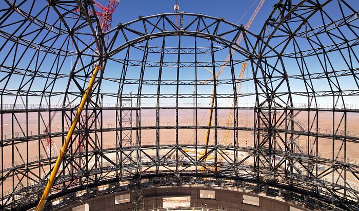This image was taken using a drone hovering in the middle of a telescope dome, under a vibrant blue sky. All around are the dark steel structures of the dome with two big arches, on the left and one at the centre. Further down is the concrete circular structure of the base. Here and there across the background, a few cranes peep out.