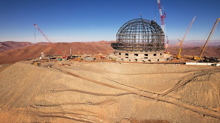 A vivid blue sky lies in the top third of this aerial image, in contrast to the orange-brown hills prominent below. The side of one hill lies in the foreground, with the criss-crossed grey steel structure of the ELT dome sitting on the flattened hilltop. More rolling red hills stretch off into the distance behind the ELT’s mountaintop.