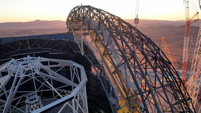 This image is centred on the grey metal framework of one of the Extremely Large Telescope's doors, which are still under construction. Also shown is a glimpse of the structure within the ELT, known as its central tower, a tall white hexagonal scaffolding. Just beyond the telescope, a golden sun sets on the dusty desert.
