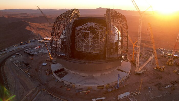 In the top right corner of the image is the sun setting over the desert mountains, casting a hue of yellow and orange over the rest of the image. In the centre of the image is the ELT, with it's white main structure surrounded by the grey dome. On either side of the opening where the main structure is are black beams that make up the arc doors.