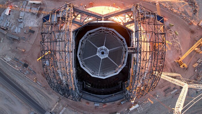 The Extremely Large Telescope sits in the very centre of this image, as seen from directly above in a photo taken by a drone. You can see right into the centre of the telescope, through the grey-panelled dome where the wide base of the central tower sits. From this angle, it looks like a giant web of hexagons that lock together to build an enormous single hexagonal unit. Either side, we see the grey metal scaffolding of the telescope's doors, still under construction. All around the telescope is the equipment of a construction site, including cranes, trucks and storage containers.