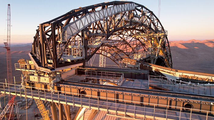 Covering the left of this image is the dull grey scaffolding of what will become one of the ELT's doors, shaped in a large arc. It is attached to a sawtooth track that spans from the bottom of the image, with one end of the door sitting on a motor control it's motion along the track. The whole image is bathed in golden sunlight from a sunset on the other side of the camera, casting long shadows against the horizon.