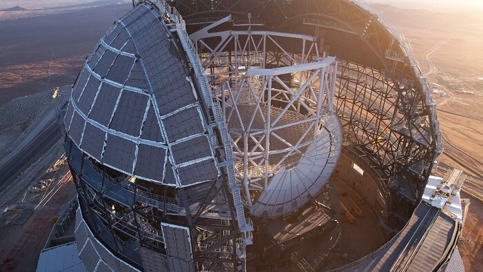 A giant dome stands in the middle of the desert. Under construction, the landscape can be seen through the frames of the dome. Inside  the dome, seen through its opening, is a giant white structure, overall cylindrical, with a honey-comb base inside. On the lateral side of the cylinder, there are a series of metallic plates that make half a circle.