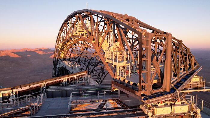 Stretching from the centre of the image to the right is the dull grey scaffolding of what will become one of the ELT's doors, shaped in a large arc. It is attached to a sawtooth track that spans from the bottom of the image, with one end of the door sitting on a motor control it's motion along the track. The whole image is bathed in golden sunlight from a sunset on the other side of the camera, casting long shadows against the horizon.