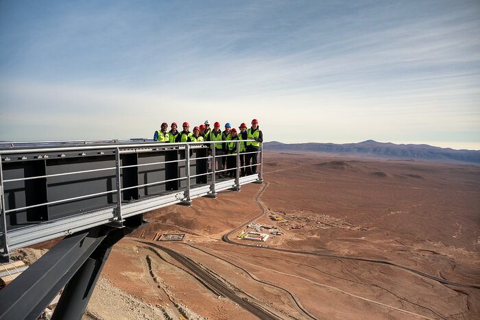 This landscape image shows a platform reaching from the left to the centre high above a brown landscape carved with lines and dotted with buildings near the lower centre. In the distance are mountains and above that a pale blue sky streaked with long white clouds. Eleven people wearing fluorescent yellow vests and hard hats of various colours stand at the end of the platform in the centre of the image.