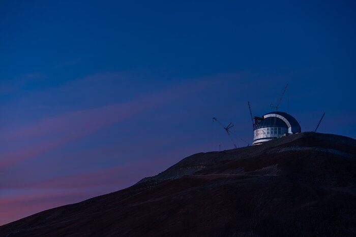 This landscape image shows a dark hill sloping down from right to left with a shiny metal structure on the top right surrounded by four cranes. The sky is blue-purple with pink-purple clouds across the centre-left of the image