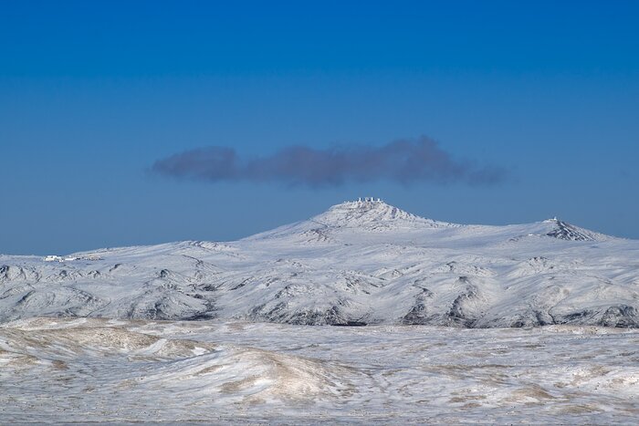 A wide view of snow-covered mountains in Chile’s Atacama Desert. Several white, rectangular telescope buildings stand along the summit of the central mountain, with a smaller structure visible on a neighbouring peak to the right, beneath a deep blue, cloudless sky.