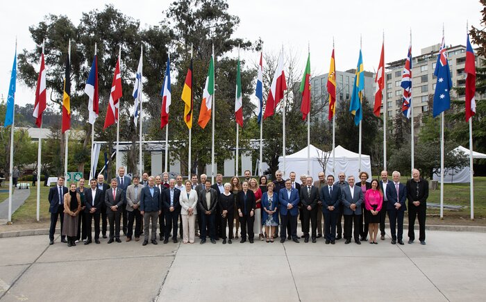 In front of several tall, hanging flags, about 40 people dressed in suits stand in two rows. A green, urban landscape fills the background.