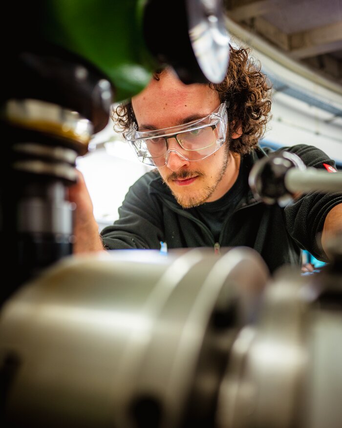 Taking up most of the image to the left and bottom is a machine, mostly metal coloured, with green details at the top. It is blurry. The focus is on a man with curly hair, operating the machine. Most of his face is visible. He is wearing a dark sweater and wearing safety glasses to protect his own glasses.