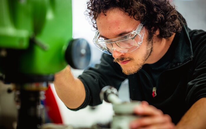 On the right, this image shows a close-up of a man with curly hair, wearing a dark sweater, operating a machine. The focus is on his face. To the left, only a part of the machine is visible, but it is blurry. The machine part is bright green and has a black knob. The man is wearing safety glasses to protect his own glasses.