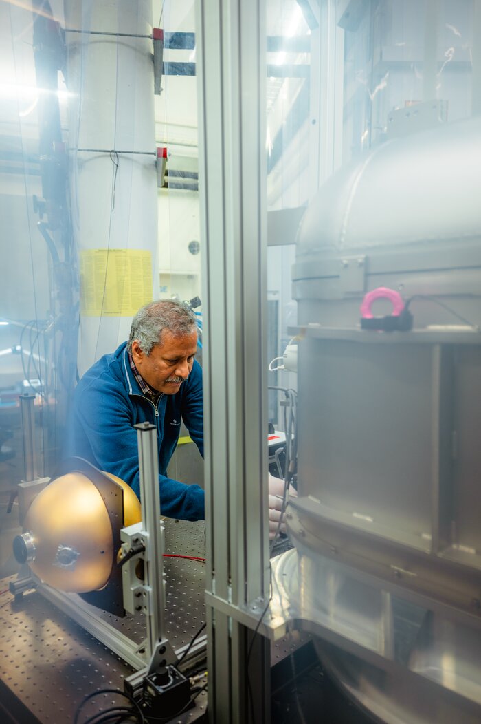 The image focuses on a man with a blue pullover working at ESO's laboratories. The laboratory is painted white, but is extremely busy with instruments and cables. The man appears handling a couple of small objects in front of a large metallic machine on the right, almost as tall as the man himself. The image is cut in the middle by a metallic column which appears to be part of the lab's infrastructure. Besides him lies a spherical golden orb bigger than a football, seemingly attached to a small reprostand.