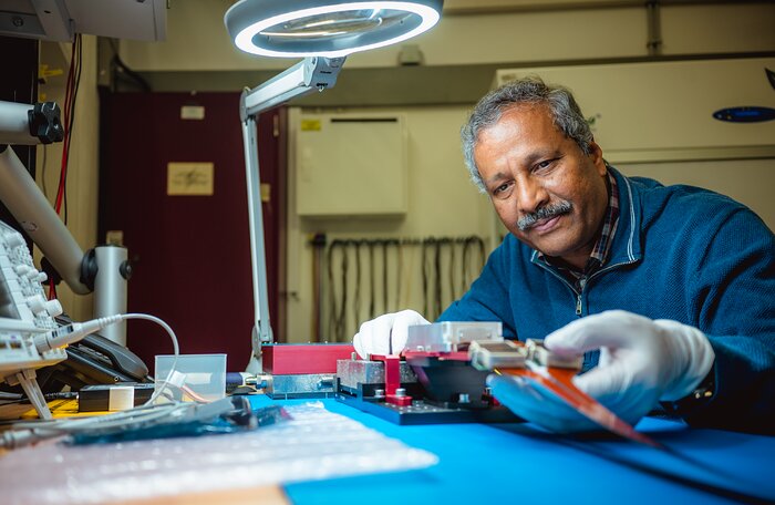 Dr. Naidu Bezawada is sitting in a closed laboratory, while inspecting and handling a black and red electronic instrument. The working table is lit by a magnifying glass as big as a hand which includes a circular LED light. Other instruments on the table include a grey squared electronic instrument with cables connected, partly visible, and a black telephone behind it.