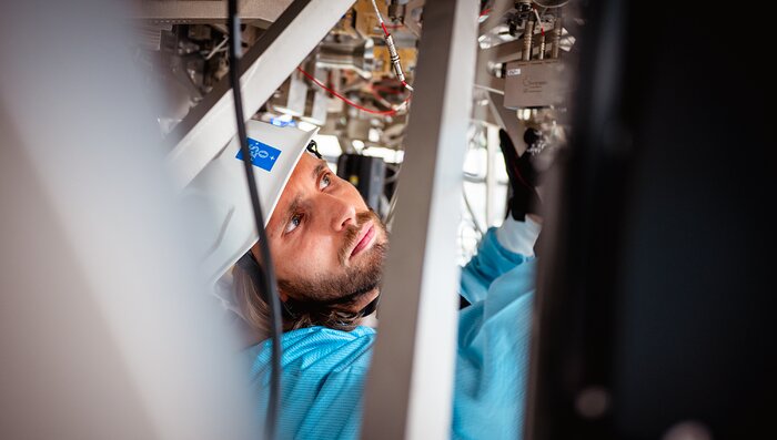 In the centre of the close-up image, Paulo Ferreira is in blue overalls with a white safety helmet, looking to the right, fitting parts to a machine. To both sides, the foreground is blurry. In the background, metal parts and cable wires are hanging above him.