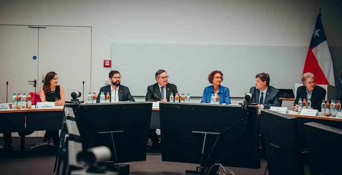 Three men and three women are sitting on the corner of a large, round, white table. On the table there are small black microphones and bottles of water and apple juice. Behind the table stands Chile's flag.