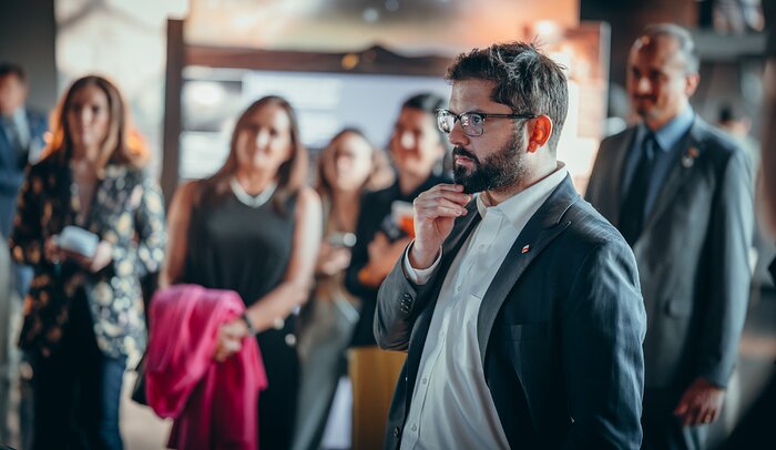 The image focuses on a man with brown short hair and short facial hair (Chile's president Gabriel Boric), wearing glasses and a dark suit with a white shirt. The president appears to be listening to someone or observing an object out of the picture's frame. On the background stand five other people, who appear blurry and are similarly looking in the same direction.