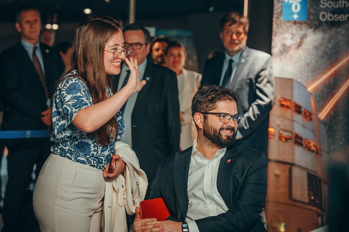 The image shows a woman with long straight to curly hair, standing, waving her hand. She is wearing glasses and a white shirt with blue flora motifs. To the right there is a man sitting down on a chair, with short, brown hair and glasses, and a short beard. Both are looking somewhere to the left of the image, smiling.