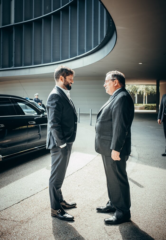 Two men stand in front of each other outside of a large black building. The man on the left, Chile's President Gabriel Boric, has short brown hair, short facial hair and is wearing glasses, a dark suit and a white shirt. To his right stands ESO Director General Xavier Barcons, who has grey hair and wears a pair of glasses together with a dark suit, a white shirt and a blue tie. There is also a black car on the background.