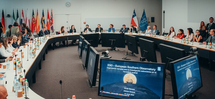 A large, donut-shaped white table occupies most of a white room. The table is occupied by dozens of people. On the table there are paper cups, bottles of water, orange juice and apple juice. In the centre there are a series of screens facing the attendees, which show a slide with the Extremely Large Telescope and a full Moon together with the letters 