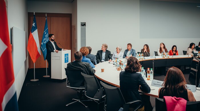 A man stands behind a podium with ESO's blue logo, delivering a speech. Besides him there is a large white table, occupied by a group of people who are looking at him. On the table, there are bottles of water, apple juice and small microphones, as well as the names of the attendees printed.