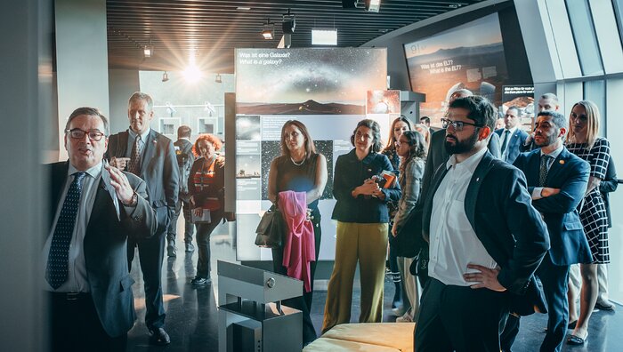 A group of people, many wearing suits and pins with the Chilean flag, are actively listening to a man on the left side of the picture. The man, ESO Director General Xavier Barcons, has brown to grey hair, wears glasses, a dark suit, a white shirt and a blue tie. The man is lifting his left hand while looking at an object outside of the picture. On the forefront, right section of the picture stands a man with short brown hair, short facial hair, wearing glasses, a dark suit and a white shirt, who is also looking at Xavier Barcons. On the background there are different panels displaying astronomical objects or views of the Atacama desert together with adjacent information.