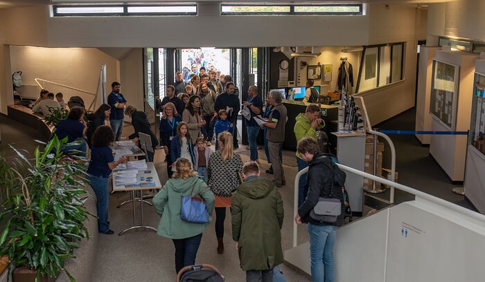 The image shows the entrance of a building. Through the glass doors it is possible to see a queue of people waiting outside. Inside the building there are already crowds engaging with volunteers (identifiable by their blue vests), as well as tables with useful information to any visitor.
