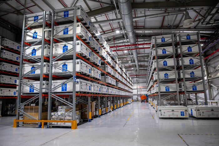 This landscape image shows a warehouse with metal structures holding white cuboid boxes, each wider than they are tall, and all with a blue ESO logo on their side. 3 racks are shown, and each holds a 2x5 cross section of boxes, with many columns down the length of each rack.