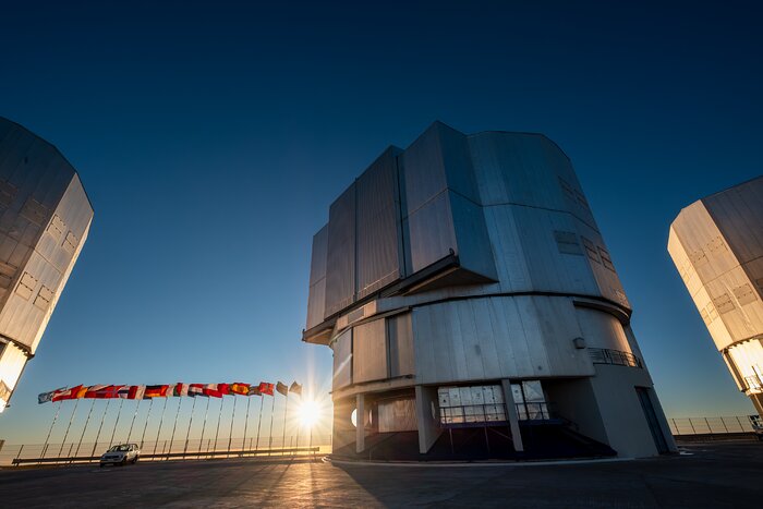 This landscape image shows a large metal structure on the centre-right, and the edges of similar metal structures on the left and right. Between the left and centre metal structures is a parked car, 18 flags flying on posts, and the Sun low in the sky. The sky is a dark blue at the top of the image and light blue to pale yellow at the bottom of the image.