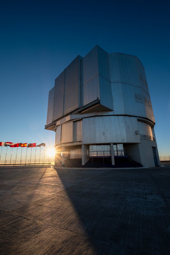 This image in portrait shows a large metal structure on the centre-right. To its left the Sun low in the sky and 10 flags are flying on posts. The sky is a dark blue at the top of the image and light blue to pale yellow at the bottom of the image.