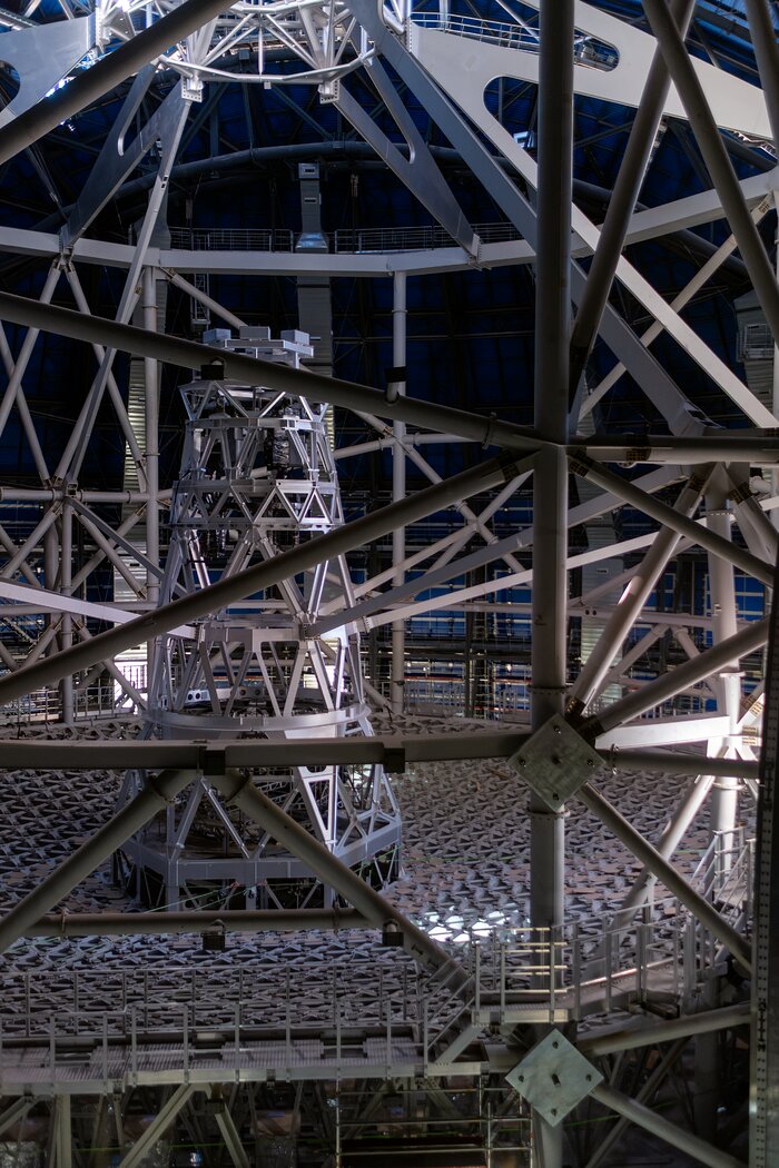 This image in portrait shows metal beams in the foreground, the plane of a metal grid in the centre, and a tall metal post in the centre of the grid and centre-right in the photograph.