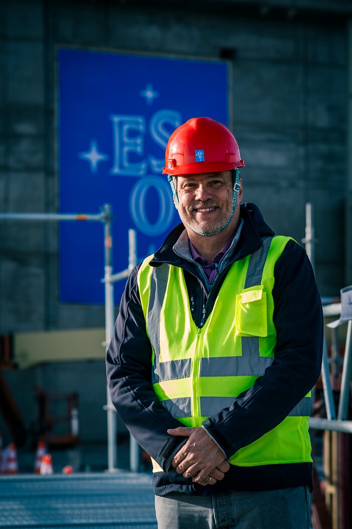 This image in portrait shows a man standing with his hands clasped in front of him. He is wearing a red hard hat and fluorescent yellow vest on top of a black jacket. Behind him is some scaffolding and a large blue sign with the ESO logo on it.