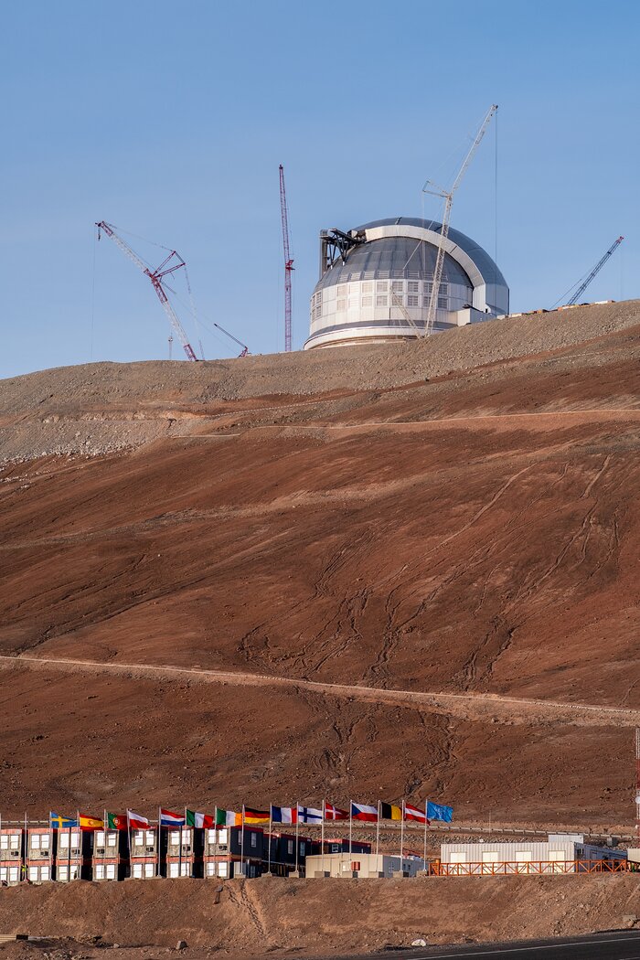 This image in portrait shows a metal structure surrounded by cranes and a light blue sky in the top third, and a red-brown hill in the lower two-thirds with paler diagonal lines in a zigzag across it. Near the bottom of the image are several buildings and in front of them15 flags are flying on posts.