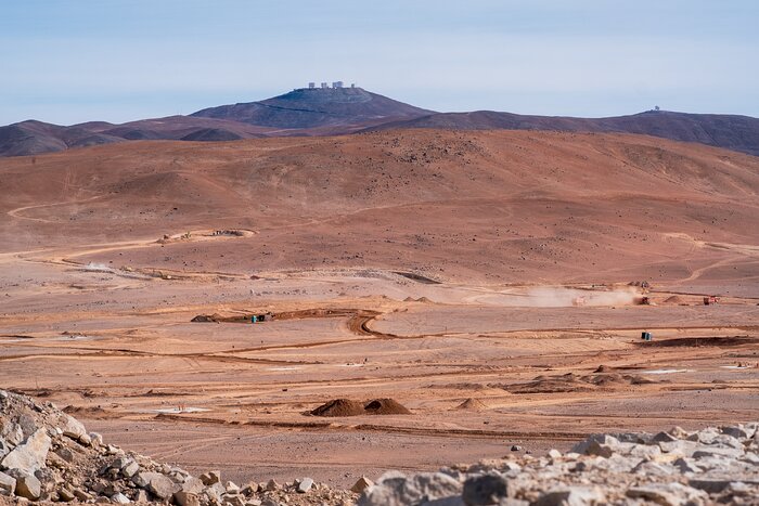 This landscape image shows a brown-orange ground in the bottom two-thirds, with darker coloured mountains in the background and rocks in the foreground. The ground is carved with lines and mounds of dirt are scattered around. Multiple large structures can be seen atop a mountain in the distance, just right of the central line, and the sky above is pale blue with horizontal white clouds.