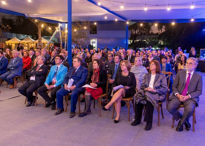 Under a large white tent roof, dozens of people dressed in suits sit in rows of chairs, looking off camera to the left. Strings of lights brighten the tent in contrast to the dark outside.