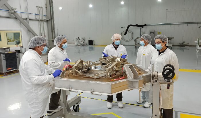 Inside a large white warehouse, five people in protective lab coats, masks, and hair nets stand around a metal, hexagonal object, suspended above the ground.