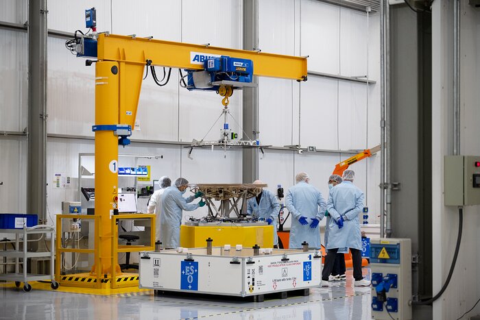 A group of people in scrubs, hair nets, face masks and gloves stand around a yellow-orange platform, on which a metallic structure rests. One person is using a tool on the structure. A yellow-orange crane hovers over it. An empty white crate sits on the floor in front of the platform; the ESO logo — four white stars surrounding the letters E, S, O over a blue background — can be seen on the crate and other parts of the room.