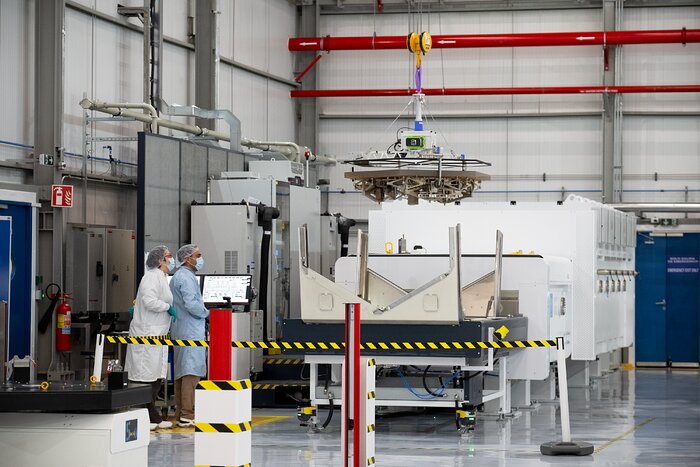 A hexagonal mirror segment is being lowered by crane into a metal container in the centre of the image. Two people in lab coats, gloves, hair nets, and face masks stand in front of computers beside the box and watch it be lowered.