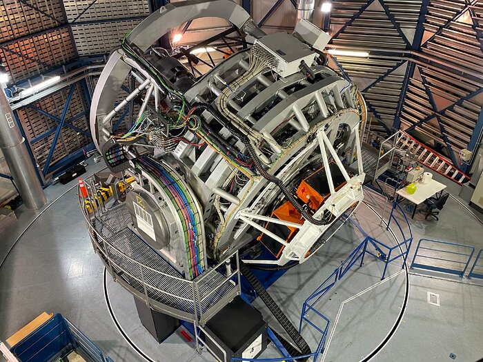 Das Foto zeigt das VISTA-Teleskop der Europäischen Südsternwarte (ESO) in seiner Kuppel am Paranal-Observatorium in Chile. Das große, silbern-graue Teleskop dominiert den Raum; es steht auf einer drehbaren Plattform und ist von Geländern, Kabeln und technischen Geräten umgeben. Deutlich sichtbar ist der neu installierte 4MOST-Spektrograf – ein komplexes Instrument mit zahlreichen bunten Kabeln, Rohren und weißen Stützstrukturen, die sich um die Rückseite des Teleskops winden. Zwei orangefarbene Elemente markieren den Bereich, in dem 4MOST befestigt ist. Im Hintergrund sind die metallisch-blauen Streben der Kuppel sowie Arbeitstische mit Werkzeugen und Computern zu sehen, die auf laufende technische Tests hindeuten. Die Szene vermittelt den Eindruck einer hochmodernen astronomischen Anlage während der erfolgreichen Inbetriebnahme eines neuen wissenschaftlichen Instruments.