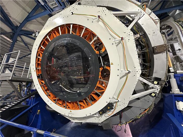 Das Foto zeigt die Rückseite des VISTA-Teleskops mit dem installierten 4MOST-Instrument. Im Mittelpunkt steht eine große, runde Öffnung, die von einer massiven, weißen Metallstruktur mit orangen Verstrebungen eingefasst ist. Dahinter sind zahlreiche dünne Glasfaserkabel zu erkennen, die in das Innere des Instruments führen. Diese Fasern leiten das Licht von astronomischen Objekten an die Spektrografen weiter. Kabel, Sensoren und Befestigungselemente verlaufen entlang der Rückseite und sind an der Struktur fixiert. Im Hintergrund ist das Gerüst der Teleskopkuppel mit blauen Stahlträgern und technischen Plattformen sichtbar. Das Bild vermittelt einen detaillierten Blick auf die komplexe technische Infrastruktur des Spektrografen.