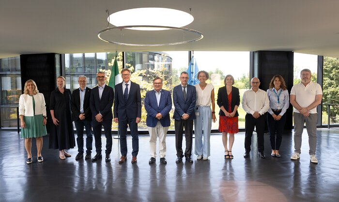 Eine Gruppe von Personen steht in einem modernen, lichtdurchfluteten Raum mit großen Fenstern und Blick ins Grüne. Die Personen sind in einer Reihe aufgestellt und blicken zur Kamera. In der Mitte tragen mehrere Anwesende Anzüge, während andere formell oder business-casual gekleidet sind. Über der Gruppe hängt ein großer, runder Deckenleuchter. Im Hintergrund ist eine Fahne mit dem ESO-Logo zu erkennen. Die Szene vermittelt den Eindruck eines offiziellen Anlasses, passend zum Kontext einer Vertragsunterzeichnung der ESO im Jahr 2026.