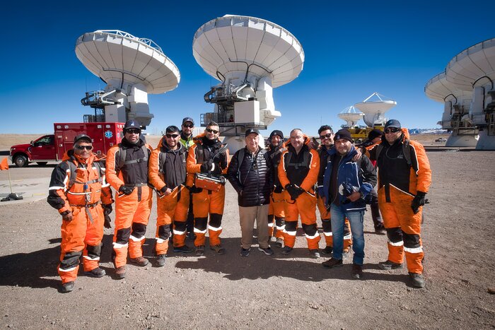 The image shows a desert landscape under blue skies, with six large, white antennas visible in the background. In the foreground, 11 people are standing in line, facing towards the camera. One person, in the centre of the image, is dressed in a black coat; of the others, nine are wearing orange work uniforms, and the final person, second-farthest to the right, is otherwise dressed.