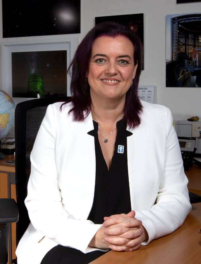 A woman is sitting down inside an office. Behind her, there is an Earth globe and different photos of astronomical objects and ESO's telescopes. The woman is dressed in a white formal jacket, wearing a pin with ESO's logo, and is smiling at the camera.