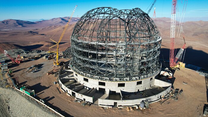 This aerial view captures almost all of the ELT construction site. The grey steel skeleton of the telescope dome sits in the centre of the image, covering about half of the scene. Below the steel, a concrete foundation rests atop the flattened mountain, with cranes and other vehicles scattered around the site. Behind the site, rust-coloured mountaintops peak above the horizon underneath a thin strip of bright blue sky.