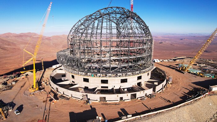 The grey steel skeleton of the ELT dome sits front and centre in this drone image. Cranes are seen to the left, the right and behind the dome structure. It rests upon a concrete base on a rust-coloured, flattened mountaintop. Behind the dome, under a bright blue sky, the rolling mountain range extends into the distance.