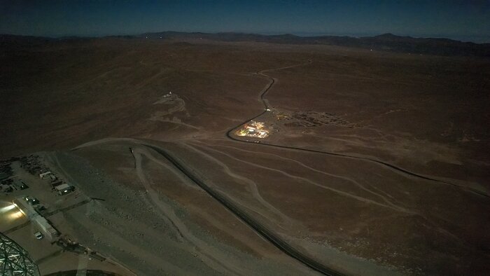 The image, taken during the night, is taken from the top of the mountain looking down. An empty brown desert stretches off into the distance, with mountaintops just visible on the horizon. The basecamp is lit up, like a small village. An access road to Cerro Armazones snakes around the basecamp and off into the distance.
