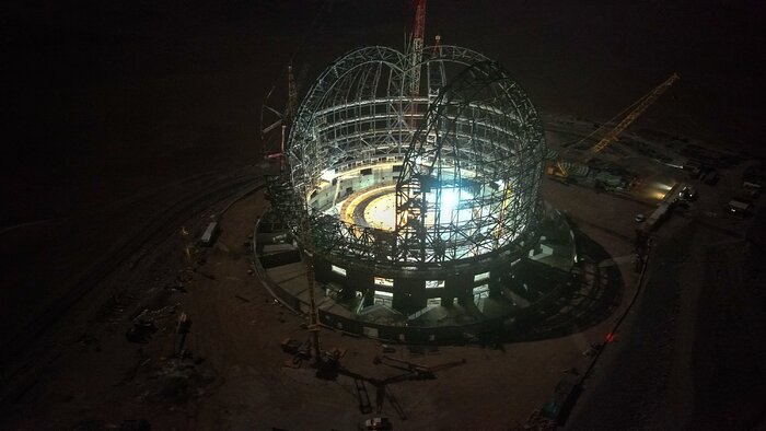 The grey steel skeleton of the ELT dome sits front and centre in this night-time aerial image. The surroundings are dark, but the faint brown hue of the ground can be seen. The inside of the dome is lit by bright blue-white lights, making the structure visible.