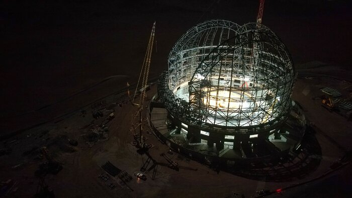 The grey steel skeleton of the ELT dome sits on the right side of the night-time aerial image. The surroundings are dark, but the faint brown hue of the ground can be seen. The inside of the dome is lit by white lights, making the structure visible.